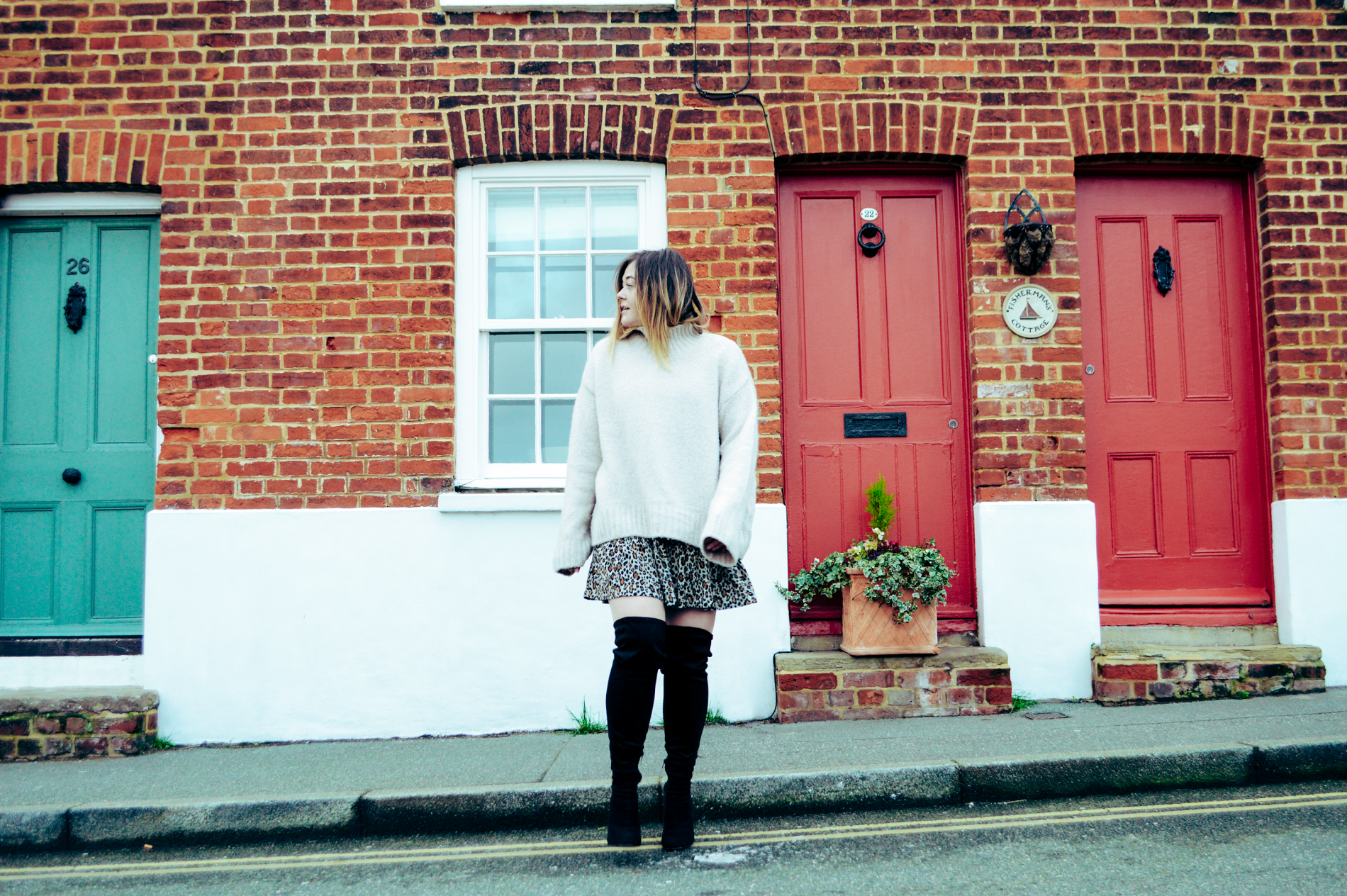 Hannah steps off of the pavement looking to the left, in front of a blue door and two red doors. Wearing a Zara leopard dress and a cream oversized jumper with over the knee black boots.